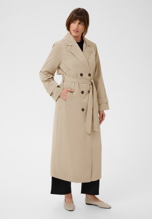 Femme portant un trench-coat beige double boutonnage avec ceinture, pantalon noir et ballerines beiges, debout avec une main dans la poche.
