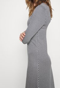 Pitkähihaisen mekon musta-valkoinen geometrinen kuosi, jossa on muotoiltu siluetti ja teksturoitu kangas. Sivuprofiilissa korostuvat muodot.