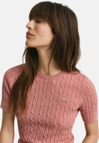 Lichtroze kabelgebreide trui met korte mouwen en een ronde hals. Bevat een klein logo op de borst. Zachte textuur.