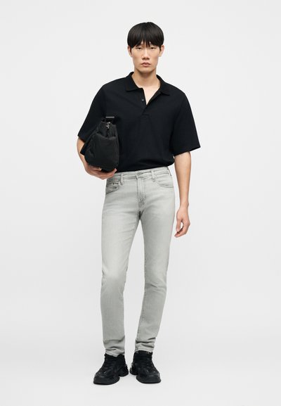 Μαύρο πόλο μπλουζάκι, ανοιχτό γκρι τζιν και μαύρα αθλητικά παπούτσια. Το μοντέλο κρατά μια μαύρη τσάντα. Απλός σχεδιασμός χωρίς ορατά σχέδια ή διακοσμητικά.