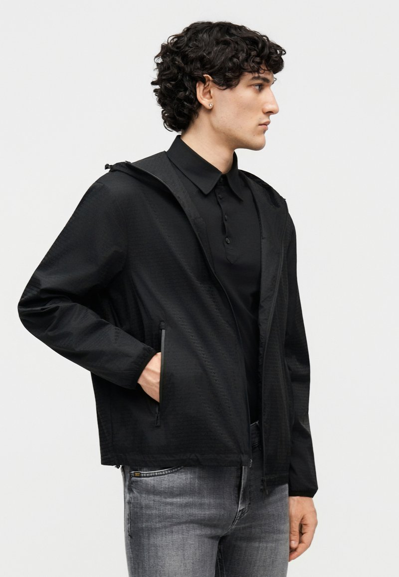 Giovane uomo con giacca nera con zip sopra una camicia nera con colletto e jeans grigi, in piedi di profilo con una mano in tasca.