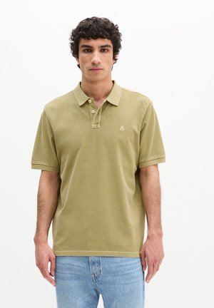 Junger Mann mit kurzen lockigen Haaren, der ein hellolivgrünes Poloshirt und hellblaue Jeans trägt, vor einem schlichten weißen Hintergrund.