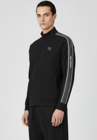 Chaqueta negra con cremallera y cuello alto, con bolsillos laterales, franjas reflectantes y un pequeño logo en el pecho. Tejido liso y texturizado.