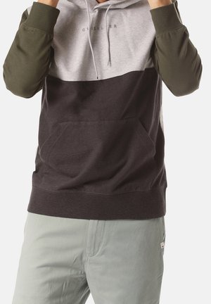 Mann trägt einen Kapuzenpullover mit Farbblock-Design: olivfarbene Ärmel, grauer Oberteil, dunkelbrauner Mittelteil und hellgraue Hose, und zieht an den Kapuzenbändern.