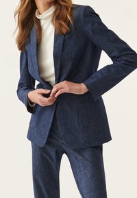Námořnický modrý upravený blazer s texturovaným povrchem, s jednou kapsou a klopovými límci, v kombinaci s odpovídajícími kalhotami.