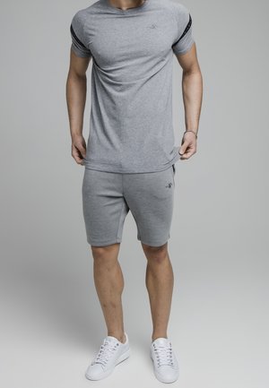 Mężczyzna w szarym, dopasowanym t-shircie z krótkim rękawem i szortach, białe sneakersy, stoi na jednolitym szarym tle.