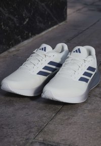 Paar weiße Sportschuhe mit marineblauen Streifen und Logos, nebeneinander auf einem strukturierten Steinboden platziert.