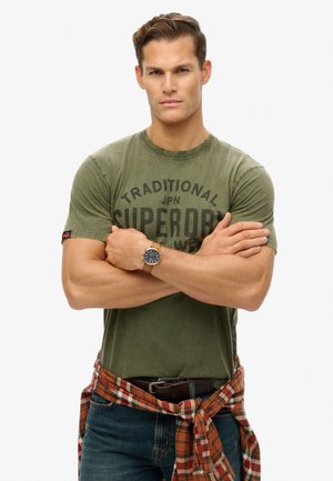 Olive green cotton T-shirt with printed text, worn with a plaid shirt tied at the waist. Features a brown watch with a blue dial.