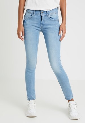 Jeans skinny de color azul claro en material de denim, con bolsillos frontales, cierre de cremallera y diseño ajustado. Combinados con zapatillas blancas.