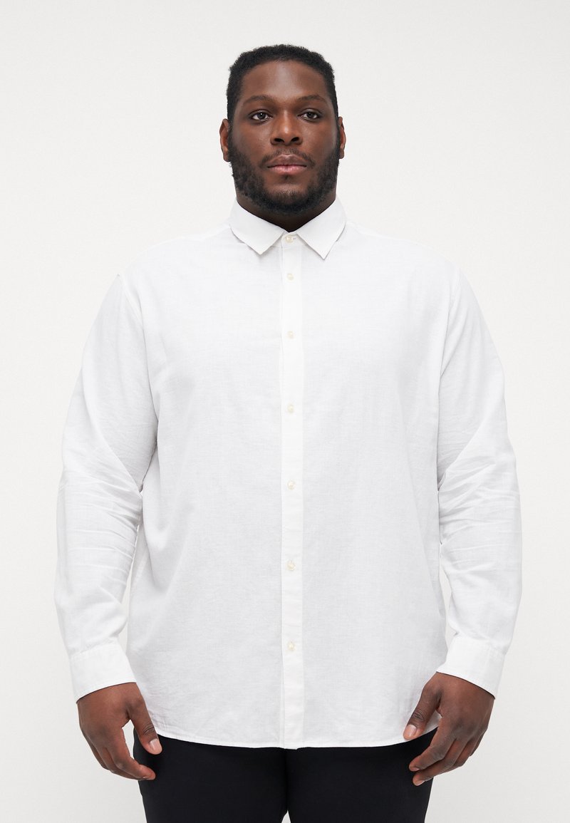 Camicia bianca a maniche lunghe con colletto classico, caratterizzata da un tessuto texturizzato e dettagli sui bottoni discreti, abbinata a pantaloni scuri.