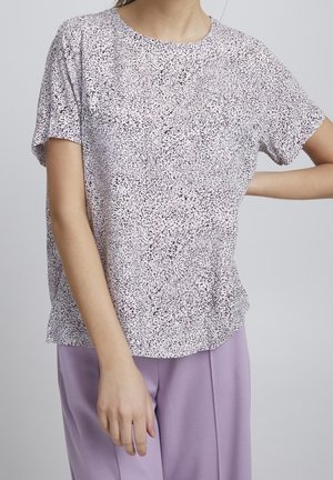 Femme portant un haut blanc à manches courtes avec des taches noires et un pantalon lavande ample, debout avec une main sur la hanche sur un fond uni.
