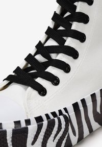 Weißes Canvas-Hochtop-Sneaker mit schwarzen Schnürsenkeln, ausgestattet mit einer kontrastierenden gummierten Sohle in schwarz-weißer Zebramusterung und strukturierten Akzenten.