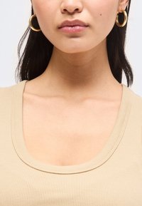 Εικόνα κοντινού πλάνου μιας γυναίκας που φορά χρυσά σκουλαρίκια κρίκους και μπεζ ριγωτό τοπ με στρογγυλή λαιμόκοψη, με μακριά σκούρα μαλλιά που φαίνονται μερικώς.