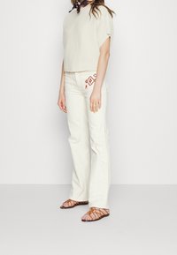 Woman wearing white textured short-sleeve top, cream pants with red geometric embroidery on pocket, brown strappy sandals, and dark scarf.