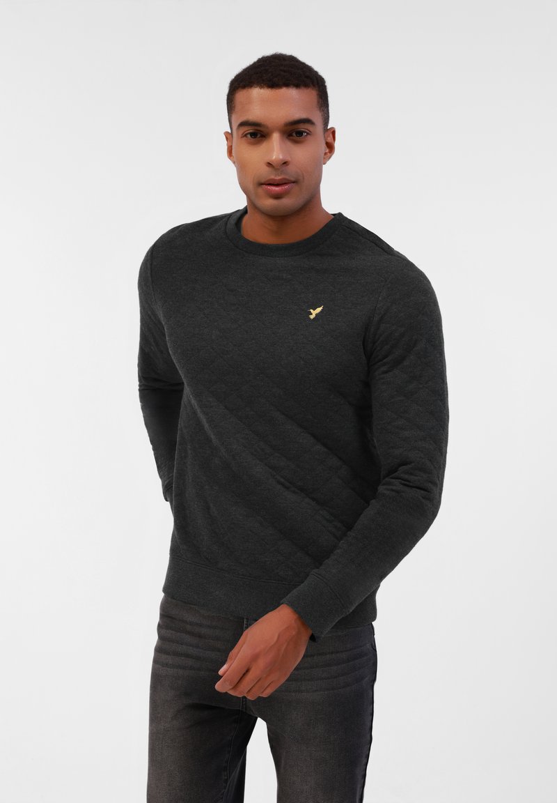 Tamno siv sweatshirt s quilted uzorkom, okruglim izrezom, rebrastim manžetama i malim zlatnim logotipom na prsima. Uparen s tamnim farmerkama za kontrast.