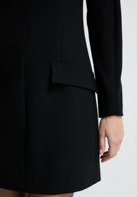 Musta muotovalettu mekko, jossa on rakenteellinen muotoilu, tasainen tasku ja sileä pinta. Pitkät hihat ja siisti, muodollinen tyyli.