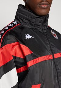 Czarna i czerwona kurtka wiatrówka z wysokim kołnierzem. Posiada biały logo Kappa, poziome i pionowe paski oraz gładką fakturę.