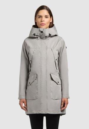 Kvinde iført lysegrå hætteparka med frontlynlås, klaplommer og justerbar hætte, stående mod en ensfarvet baggrund.