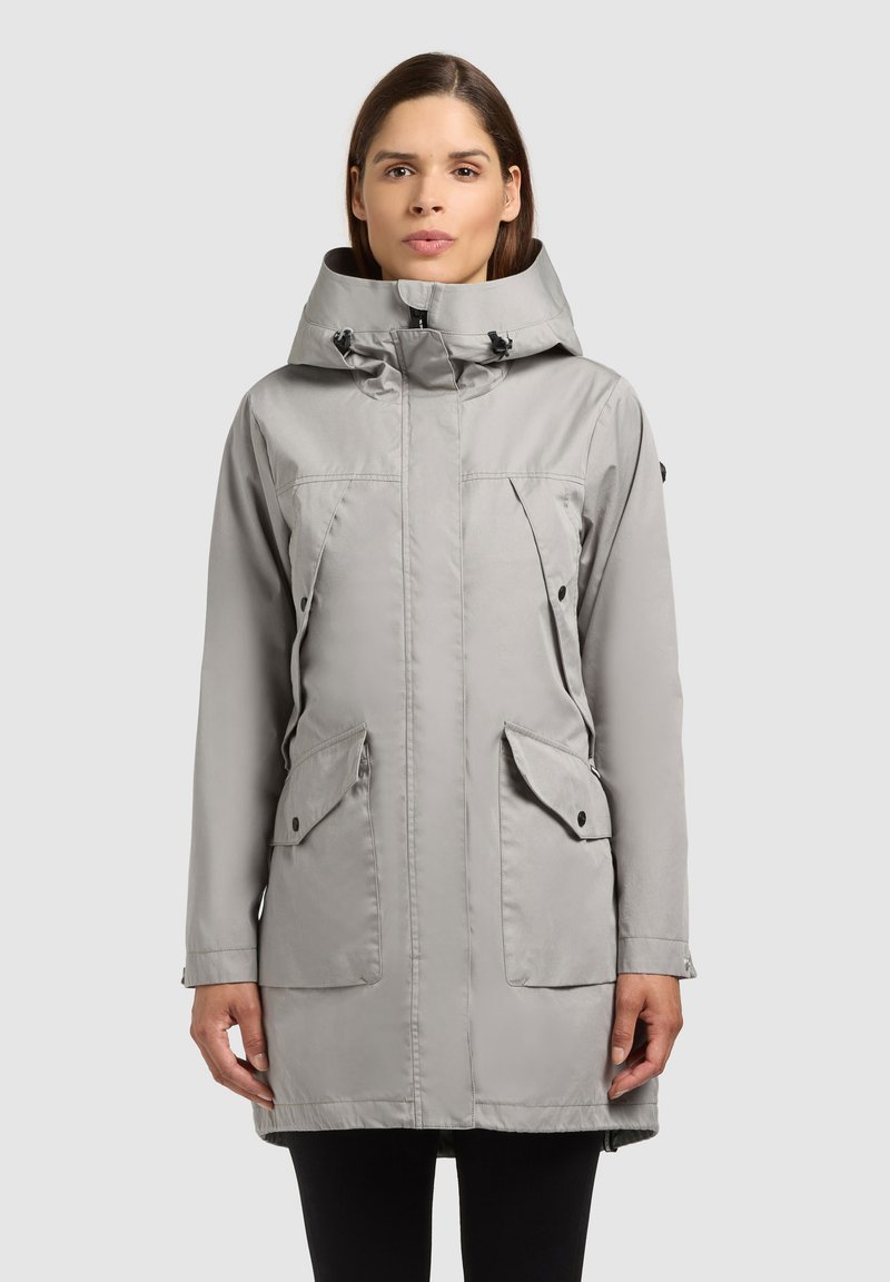 Femme portant un parka à capuche gris clair avec fermeture éclair frontale, poches à rabat et capuche réglable, debout devant un fond uni.