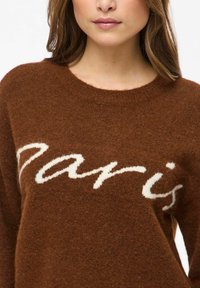 Rudas megztinis iš minkštos, tekstūruotos medžiagos su apvalia iškirpte. Priekyje baltomis raidėmis parašytas žodis „Paris“.