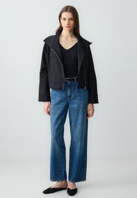 Zwarte cropped jas met een brede kraag, gecombineerd met hoge, blauwe wijde broek. Model draagt zwarte ballerina's en een nauwsluitend zwart topje.