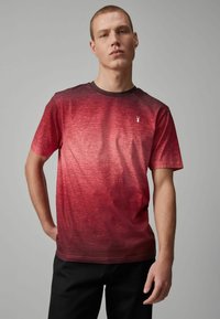 Camiseta de manga corta en degradado rojo con cuello redondo, hecha de tela texturada. Presenta un pequeño logo blanco en el pecho.