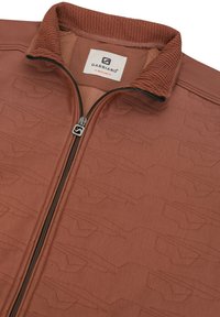 Ženska zip-up jakna v rjavi barvi, ki ima teksturirano tkanino, visok ovratnik in subtilne geometrijske vzorce. Oznaka logotipa je vidna znotraj ovratnika.