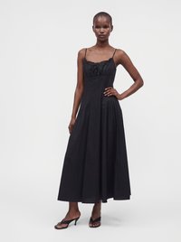 Grande femme portant une robe midi noire sans manches avec de fines bretelles et des sandales à talons noires à bout ouvert, debout avec une main sur la hanche.