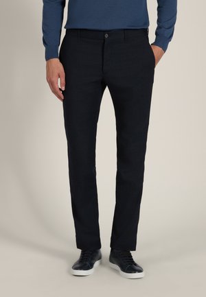 Homme portant un pantalon noir slim texturé, des baskets noires à semelles blanches et une chemise bleue à manches longues, debout avec les mains près des poches.
