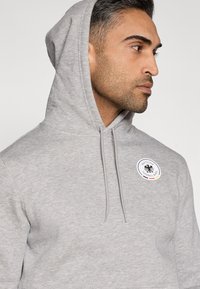 Hall halli kapuutsiga dressipluus, millel on nöör ja ümmargune plaat musta kotka logoga ning tekstiga "DEUTSCHER FUSSBALL BUND". Pehme, tekstuuriga kangas.