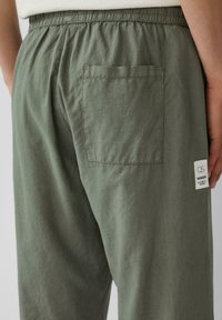 Pantalons légers gris-vert avec une taille élastique, une poche arrière et une étiquette de marque ; tissu texturé avec un léger éclat.