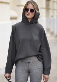 Sivi kapuljača s slobodnim krojem, dugim rukavima i rebrastim manžetama. U paru s teksturiranim sivim tajicama. Model drži sunčane naočale.