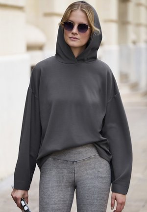 Sivi kapuljača s slobodnim krojem, dugim rukavima i rebrastim manžetama. U paru s teksturiranim sivim tajicama. Model drži sunčane naočale.