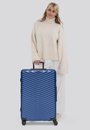Donna in un maglione beige a collo alto sta tenendo il manico di una grande valigia rigida blu con ruote su sfondo grigio.