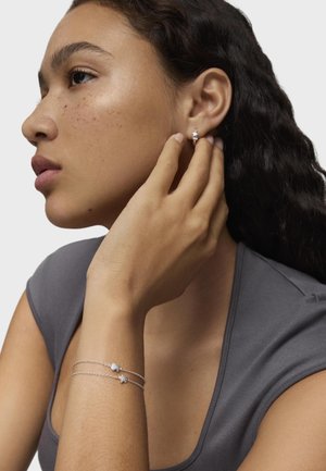 Pendientes de plata con un diseño de perla y dos pulseras de plata con charms abstractos, usados por una persona con cabello ondulado y una camiseta gris.