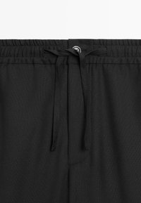 Ceinture d'un pantalon en tissu noir avec bande élastique, fermeture par bouton et cordon de serrage noué au centre devant.