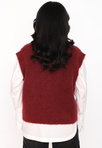 Rød, fluffy strikket utenermet genser med løs passform, båret over en hvit skjorte med krage. Har rund hals og rå kanter.