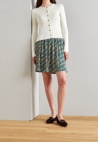 Biały prążkowany kardigan zapinany na guziki z przodu, zestawiony z wzorzystą spódnicą w odcieniach morskim, złotym i kremowym oraz czarnymi mokasynami zamszowymi ze złotymi akcentami.