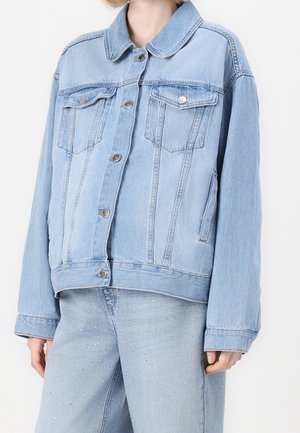 Personne portant une veste en jean bleu clair avec des boutons argentés et un pantalon en jean bleu clair assorti avec de petites embellissements.