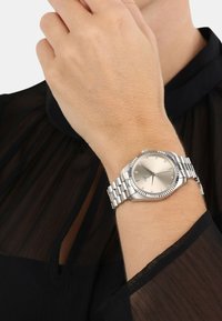 Silberner Edelstahl-Armbanduhr mit strukturiertem Lünette, einem metalischen Ziffernblatt und einem Gliederarmband. Mit Diamant-Stundenmarkierungen und blauen Zeigern.