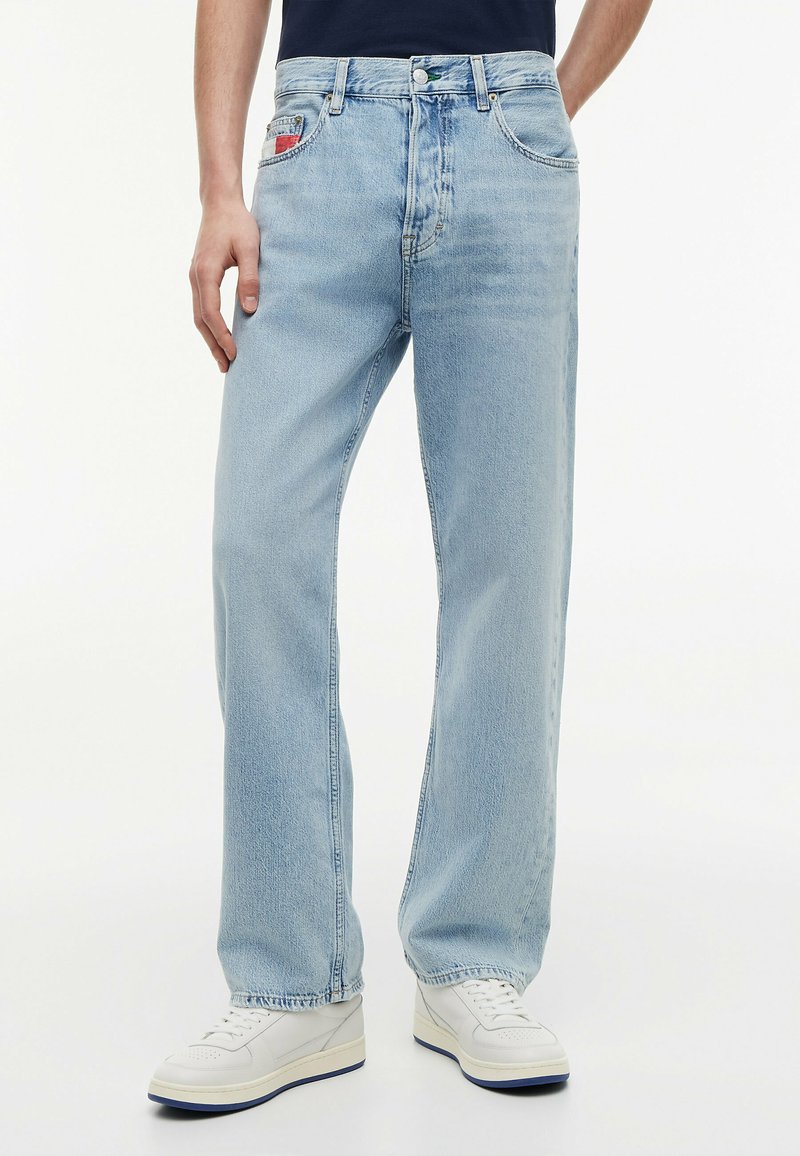 Άνδρας με ανοιχτό μπλε τζιν με ίσια πόδια και λευκά αθλητικά παπούτσια, με το ένα χέρι να ακουμπά στο πλάι, σε απλό φόντο.