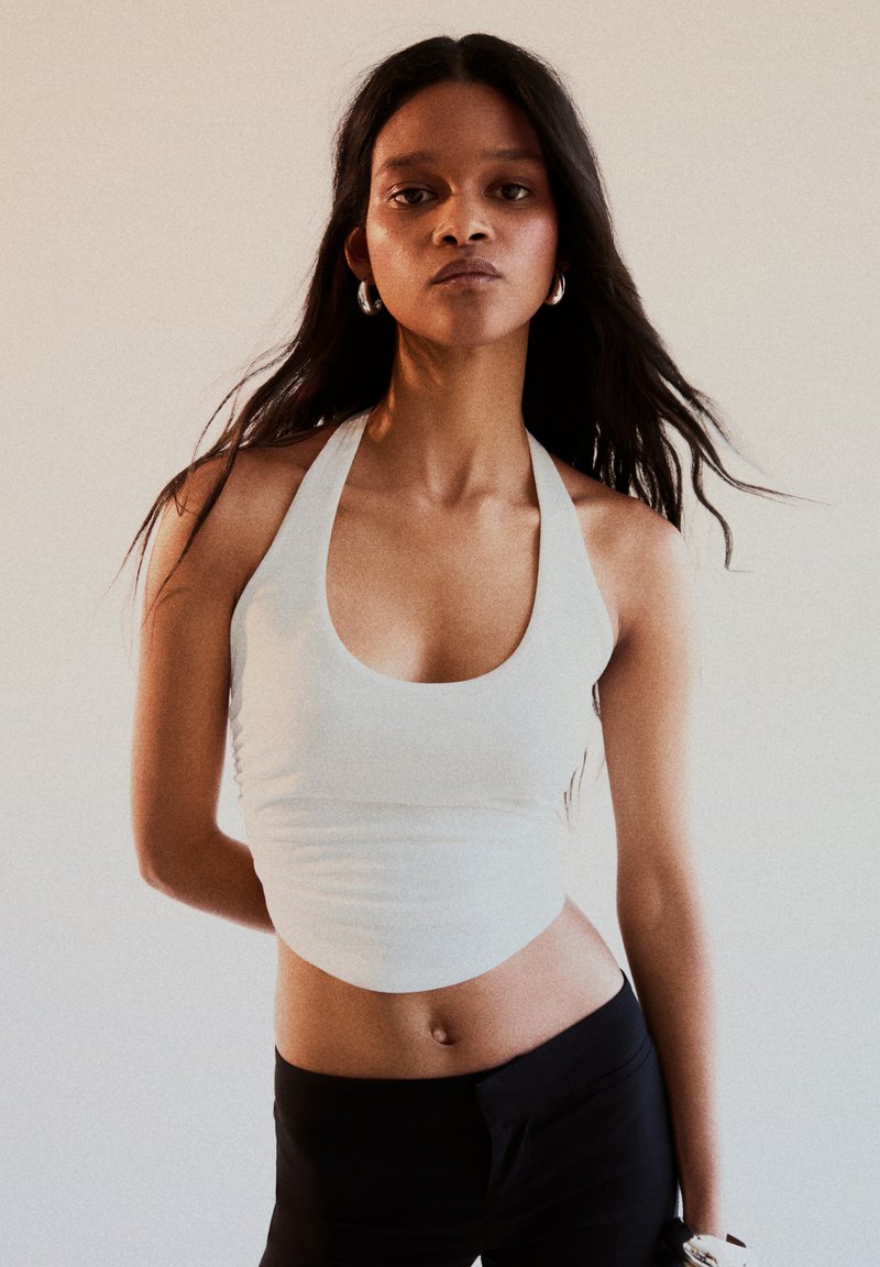 Femme aux longs cheveux foncés portant un haut blanc à col bénitier et un pantalon noir, debout avec une expression neutre devant un fond uni.