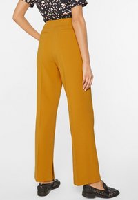 Pantalones de pierna ancha en color mostaza, hechos de una tela suave. Presentan dos bolsillos traseros y aberturas en el dobladillo. Combinados con zapatos negros.