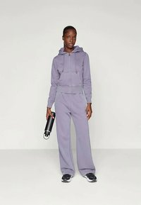 Gaiši purpursarkans īsais džemperis ar rāvējslēdzēju un regulējamu auklu, savienots ar plašām treniņbiksēm. Modelis tur melnu ūdens pudeli.