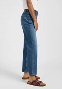 Jeans de mezclilla azul de tiro alto con corte recto, diseño clásico de cinco bolsillos y detalles de costura sutiles. Se llevan con sandalias planas.