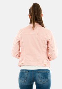 Chaqueta de mezclilla rosa con cuello, bolsillos laterales y botones dorados. Llevada sobre una camiseta blanca y combinada con jeans azules.