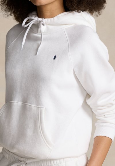 Femme portant un sweat à capuche blanc avec poche avant et petit logo brodé bleu marine sur la poitrine, manches retroussées.