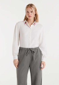 Joven mujer que lleva una blusa blanca con flores rosas bordadas en el cuello y unos pantalones de tiro ajustado a rayas grises.