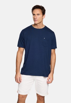 Hombre con camiseta de manga corta azul marino con bolsillo en el pecho y pantalones cortos blancos remangados, de pie contra un fondo claro y liso.