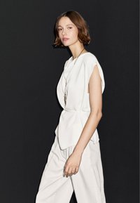 Hvid peplum top med kapærmer; blødt stof, glat tekstur. Sammen med løstsiddende off-white bukser, minimal detaljer. Mørk baggrund.